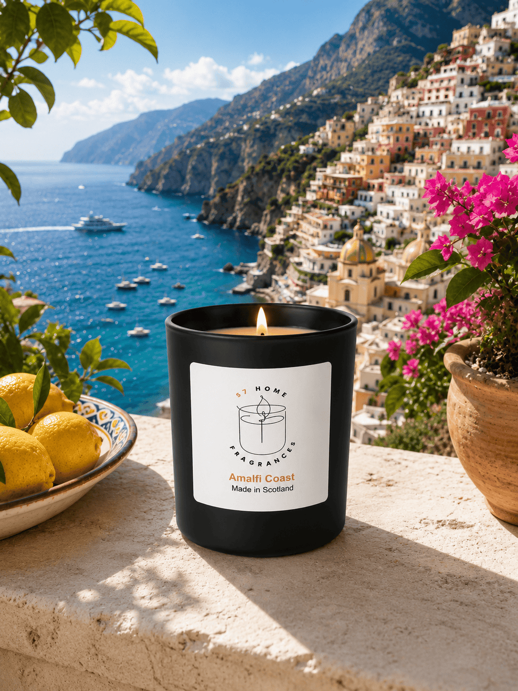 A black scented candle jar with a white label reading 'Amalfi Coast' and 'Made in Scotland' sits on a stone ledge overlooking a scenic coastal town and blue sea. Yellow flowers and lemons are visible in the foreground.