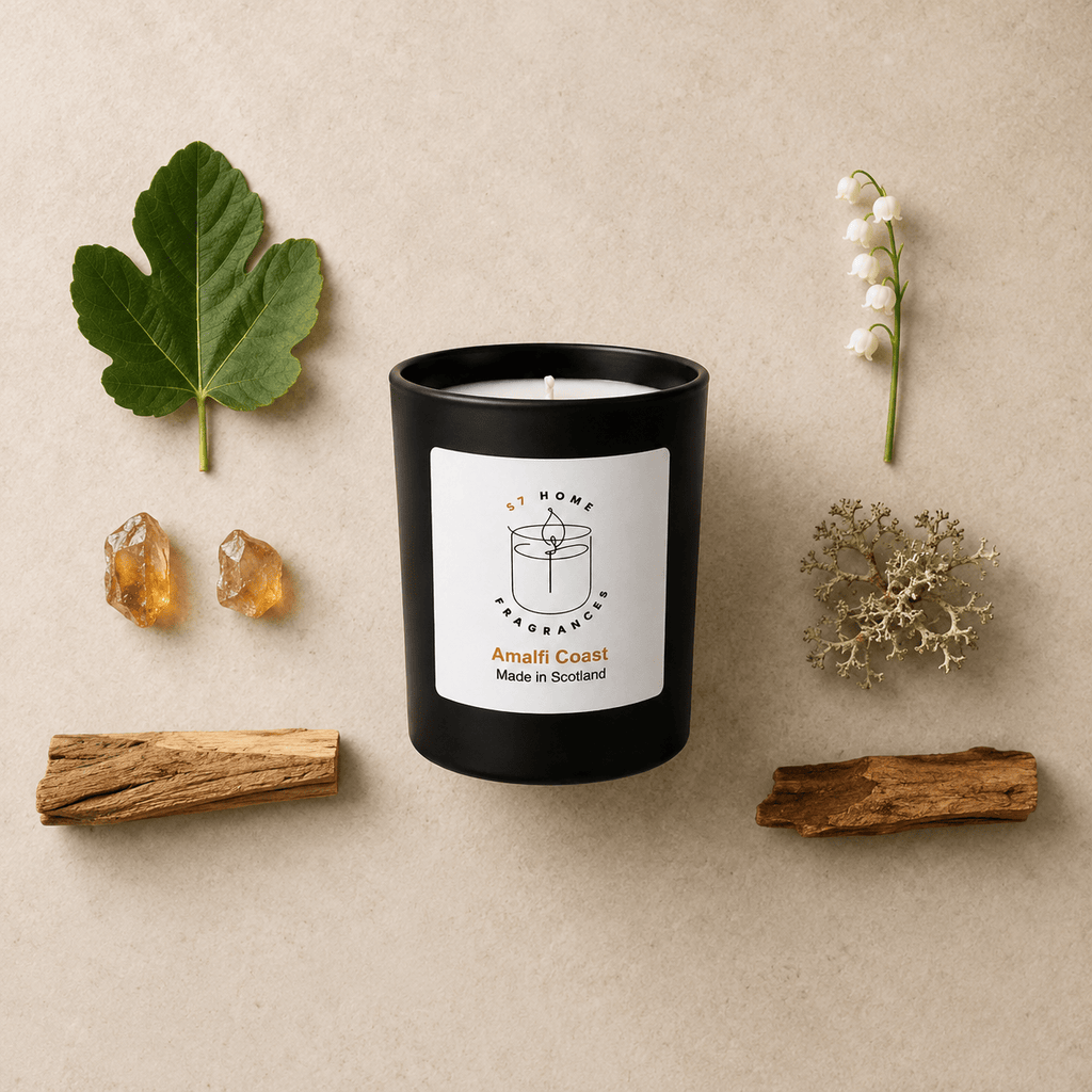 A black glass candle jar with a white label reading 'S7 Home Fragrances' and 'Amalfi Coast Made in Scotland,' styled on a beige surface with a green leaf, two amber stones, a white flower stem, dried botanicals, and two pieces of wood.