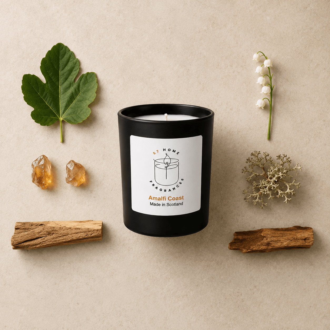 A black glass candle jar with a white label reading 'S7 Home Fragrances' and 'Amalfi Coast Made in Scotland,' styled on a beige surface with a green leaf, two amber stones, a white flower stem, dried botanicals, and two pieces of wood.