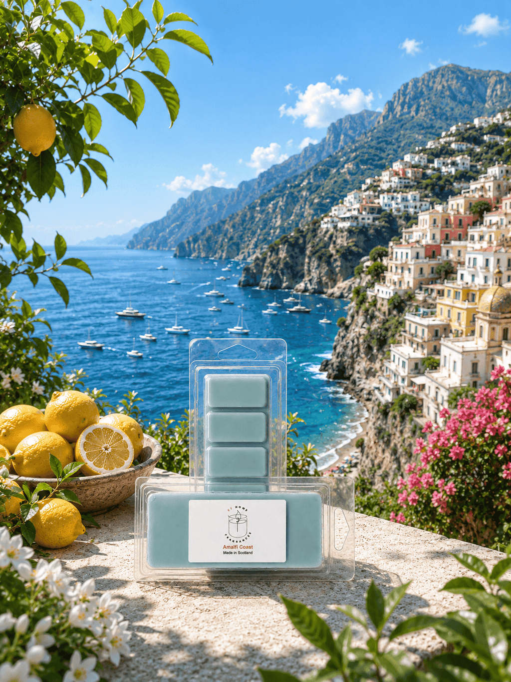A packaged bar of a wax melt product labeled 'Amalfi Coast' is displayed in front of a scenic coastal landscape with blue sea, mountains, and lemon trees. The product itself is pale mint green and sits on a stone surface with lemons and flowers around it.