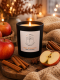 Candle labeled 'Apple & Cinnamon' surrounded by apples and cinnamon sticks on a wooden surface.