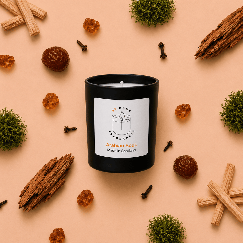 A black scented candle jar with a white label reading 'S7 Home Fragrance' and 'Arabian Souk Made in Scotland,' arranged on a peach background with cinnamon sticks, cloves, amber pieces, moss, and bark around it.