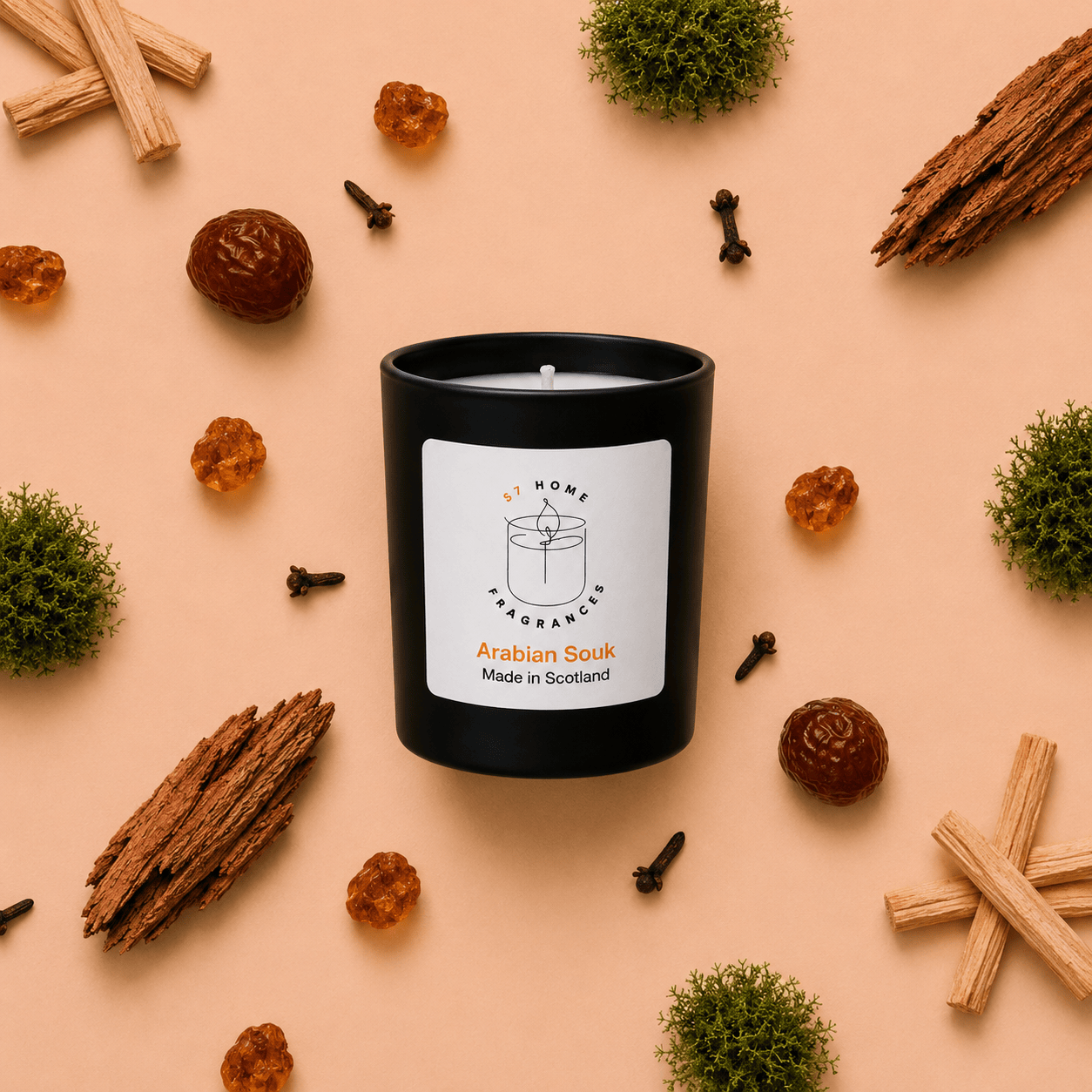 A black scented candle jar with a white label reading 'S7 Home Fragrance' and 'Arabian Souk Made in Scotland,' arranged on a peach background with cinnamon sticks, cloves, amber pieces, moss, and bark around it.