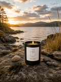A black glass jar candle with a white label reading 'Argyll Shores' and 'Handmade in Scotland' sits on a rocky shoreline at sunset, with water, mountains, and grasses in the background.