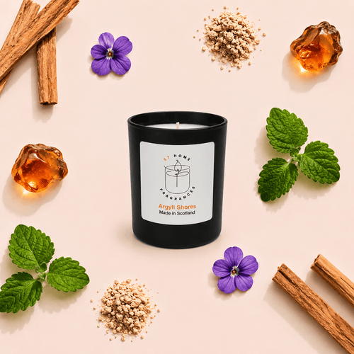 A black glass candle jar with a white label reading 'S7 Home Fragrance' and 'Argyll Shores Made in Scotland,' styled on a pale pink background with cinnamon sticks, purple flowers, mint leaves, and amber cubes around it.