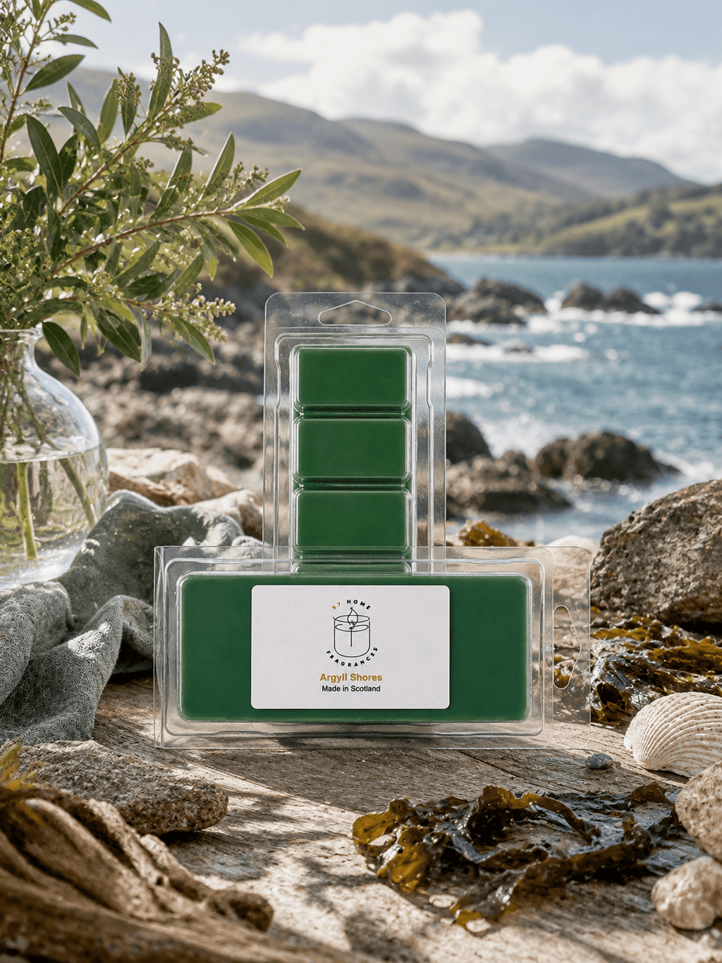 A packaged set of green wax melts or scented melts displayed on a rocky beach with ocean, mountains, rocks, and greenery in the background. The front label reads 'Argyll Shores' and 'Made in Scotland.