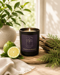 A lit black glass candle jar with a dark label reading 'S7 Home Fragrances' with a scent of 'Bergamot & Cedarwood' sits on a wooden board in a styled setting with a lime, greenery, and a vase in the background.