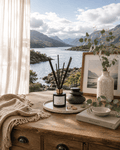 A reed diffuser with black reeds in a black glass bottle and gold collar sits on a rustic wooden table in a styled interior. The scene includes a cream throw blanket, a ceramic vase with greenery, a framed print, a small bowl, and a book, all set against a large window overlooking a scenic mountain lake. Scent is Caledonian Breeze, made in Scotland by S7 Home Fragrances