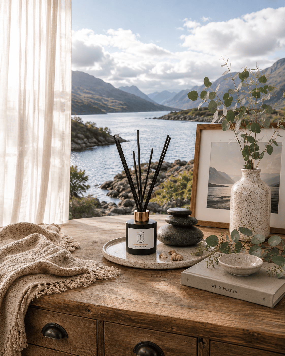 A reed diffuser with black reeds in a black glass bottle and gold collar sits on a rustic wooden table in a styled interior. The scene includes a cream throw blanket, a ceramic vase with greenery, a framed print, a small bowl, and a book, all set against a large window overlooking a scenic mountain lake. Scent is Caledonian Breeze, made in Scotland by S7 Home Fragrances