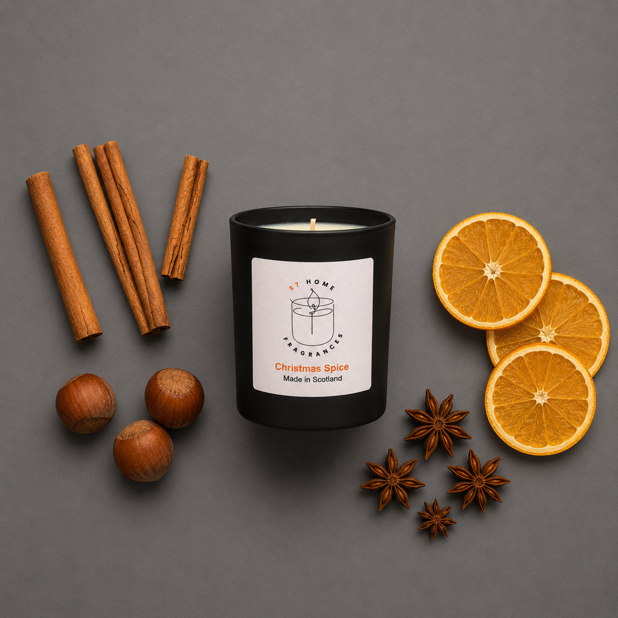 Black candle labeled 'Christmas Spice' surrounded by cinnamon sticks, oranges, and nuts on a gray background