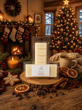 A packaged set of cream-colored wax melts displayed on a rustic wooden surface in a cozy holiday room. The scene includes a lit fireplace, stockings, a Christmas tree with warm lights, a mug, dried orange slices, cinnamon sticks, and decorative cookies.