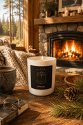 A white candle with a black label reading 'S7 Home Fragrances' and 'Fireside' sits on a rustic wooden table in a cozy living room setting with a burning fireplace, pine needles, a book, and a mug.
