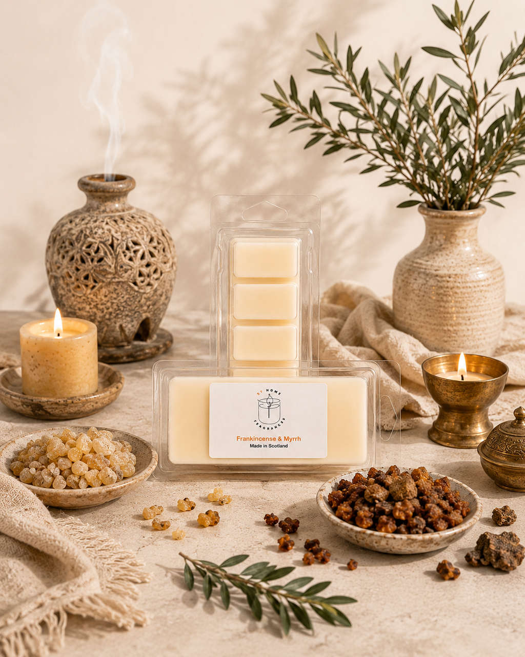 A packaged set of five cream-colored wax melts displayed in a clear plastic case, surrounded by decorative props including lit candles, a textured vase with greenery, bowls of botanical ingredients, and a small brass container on a neutral fabric surface.