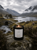 A black candle jar with a white label reading 'Highland Mist' sits on a mossy rock in a mountain landscape with a lake, bridge, and cloudy sky.
