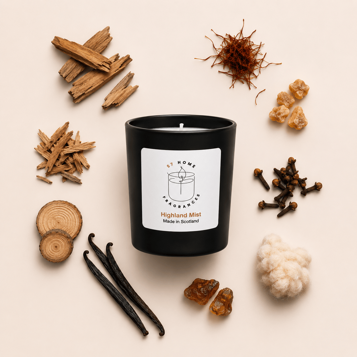 A black candle jar labeled 'A Home Fragrance Highland Mist Made in Scotland' is arranged in a styled flat lay with natural ingredients including wood slices, vanilla beans, cloves, resin pieces, and cotton fiber on a light beige background.