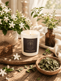 A white scented candle in a glass jar sits on a rustic wooden tray, surrounded by white flowers, green leaves, a small wooden box, and dried herbs in a warm, softly lit setting. The candle label reads 'S7 Home Fragrances' and 'Jasmine & Patchouli'.