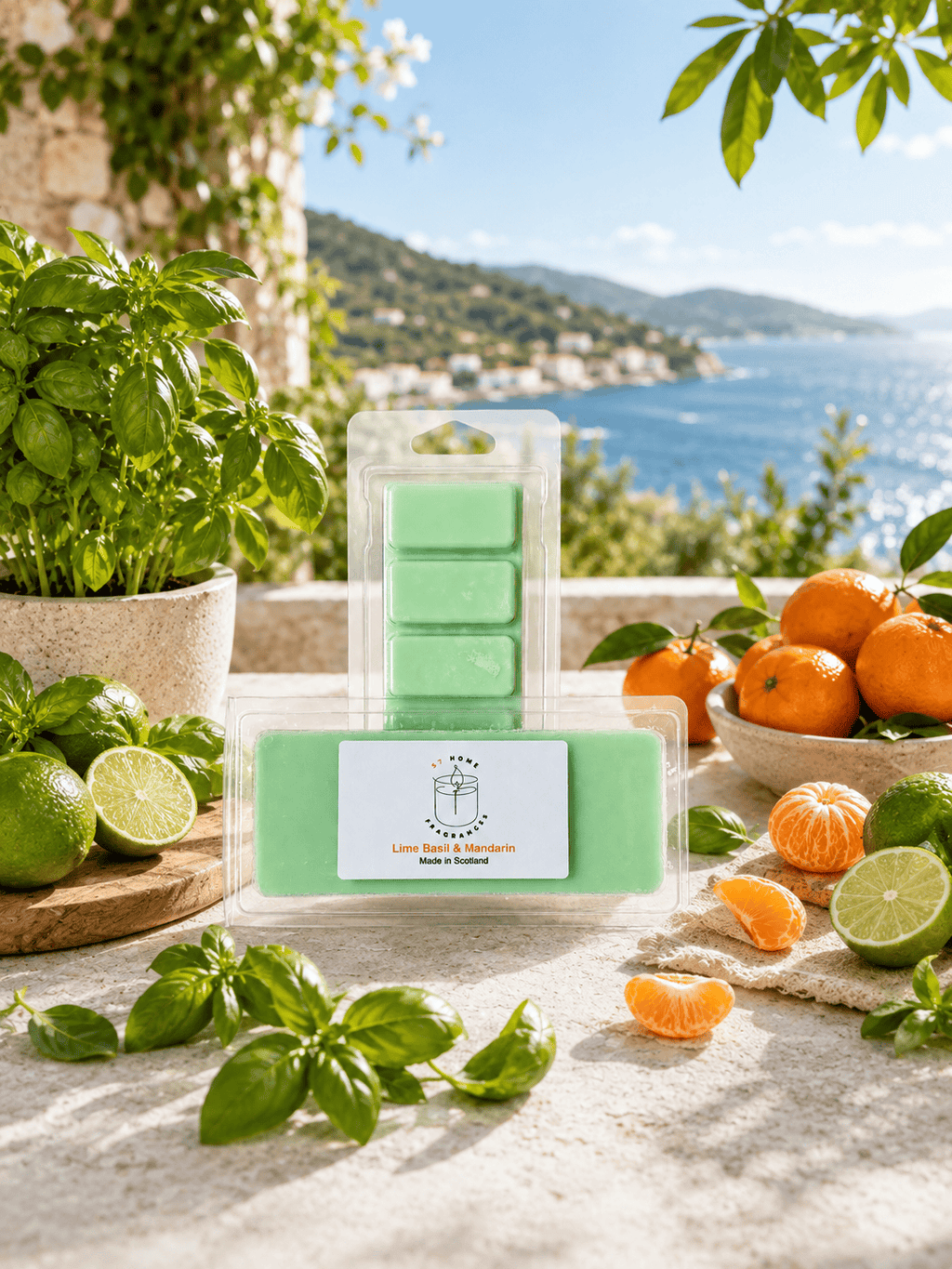 A packaged set of mint green wax melts displayed on a stone surface outdoors, with basil leaves, limes, oranges, and a scenic sea-and-mountains background visible.