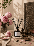 A reed diffuser bottle with black reeds and a gold collar sits on a tabletop in a styled interior scene with pink flowers, books, a brass incense burner, and warm sunlight and shadows. Scent is Peony & Oud made in Scotland by S7 Home Fragrances