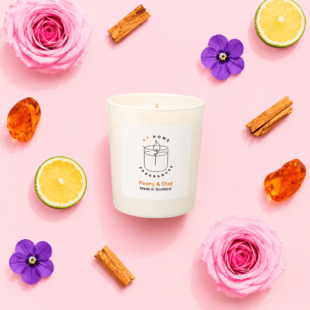 A white candle jar labeled 'S7 Home Fragrance Peony & Oud Made in Scotland' arranged on a pink background with pink roses, purple flowers, cinnamon sticks, lime slices, and amber stones around it.