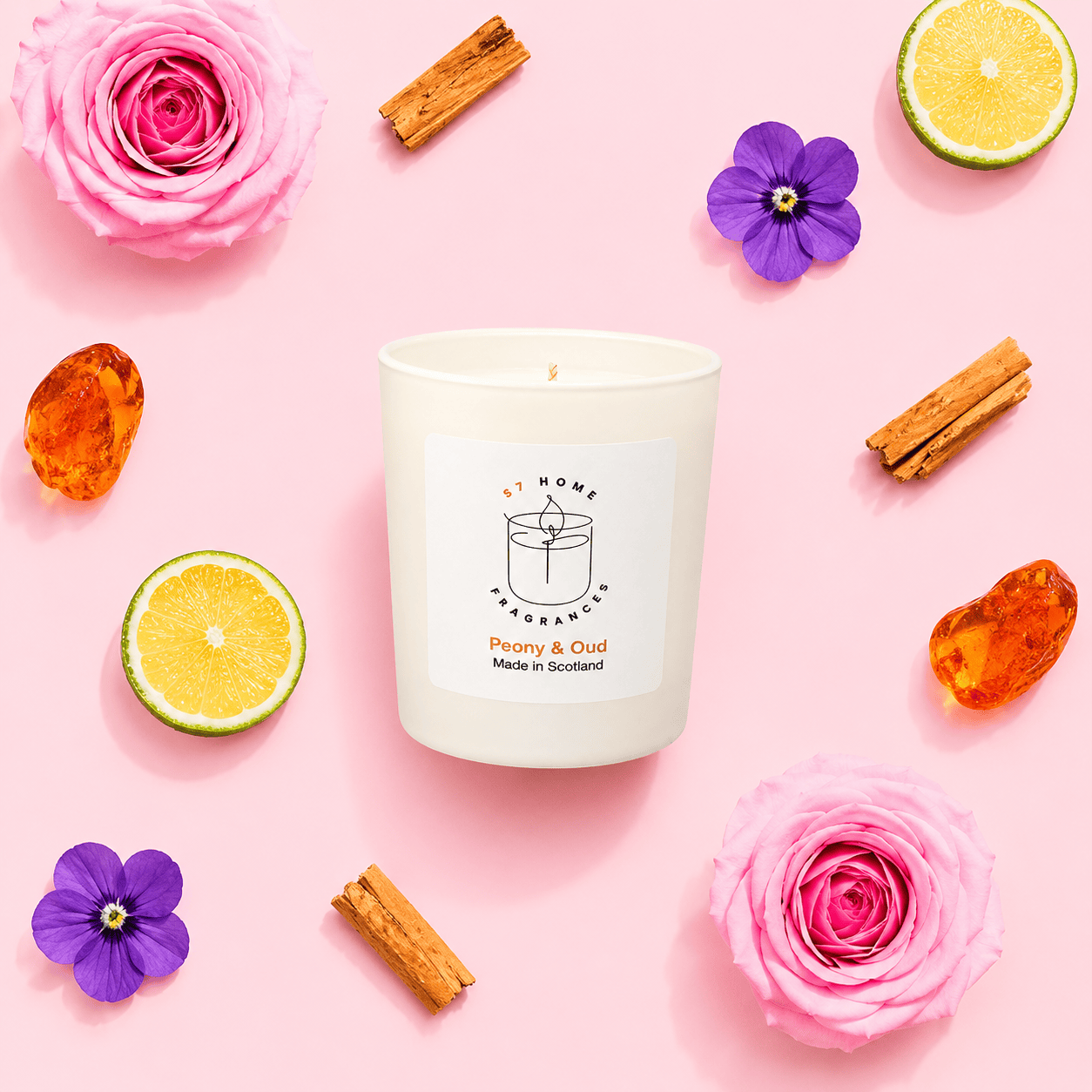 A white candle jar labeled 'S7 Home Fragrance Peony & Oud Made in Scotland' arranged on a pink background with pink roses, purple flowers, cinnamon sticks, lime slices, and amber stones around it.