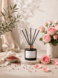 A reed diffuser bottle with black reeds and a gold collar sits on a stone slab in a styled setting with pink roses, scattered peppercorns, and draped fabric. A white label on the front reads 'S7 Home Fragrance' and 'Pink Pepper & Rose'.