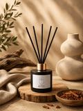 A reed diffuser bottle with black reeds, a black glass body, and a gold collar, set on a wooden slice with a beige label. The scene is styled with a beige ceramic vase, a small bowl of cinnamon sticks, and draped fabric in a warm, sunlit setting. Sandalwood scent made in Scotland by S7 Home Fragrances