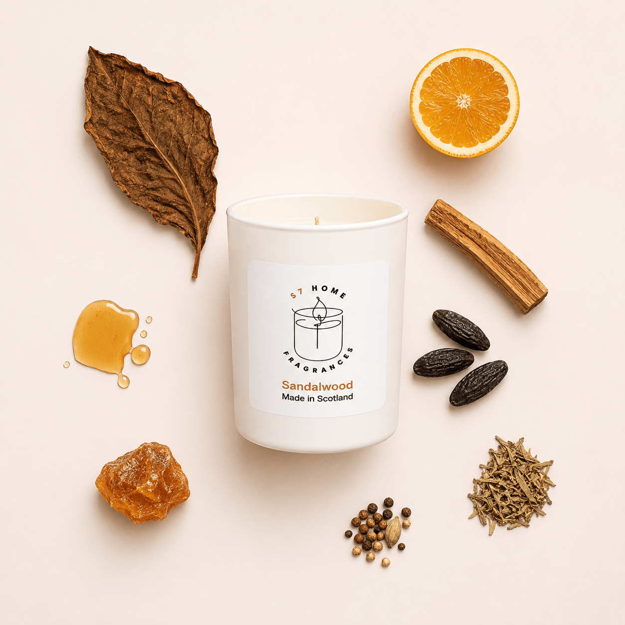A white glass candle jar labeled 'by S7 HOME Fragrances Sandalwood Made in Scotland' is arranged on a pale pink background with scent ingredients around it, including a dried leaf, orange slice, cinnamon stick, honey drop, seeds, and spices.