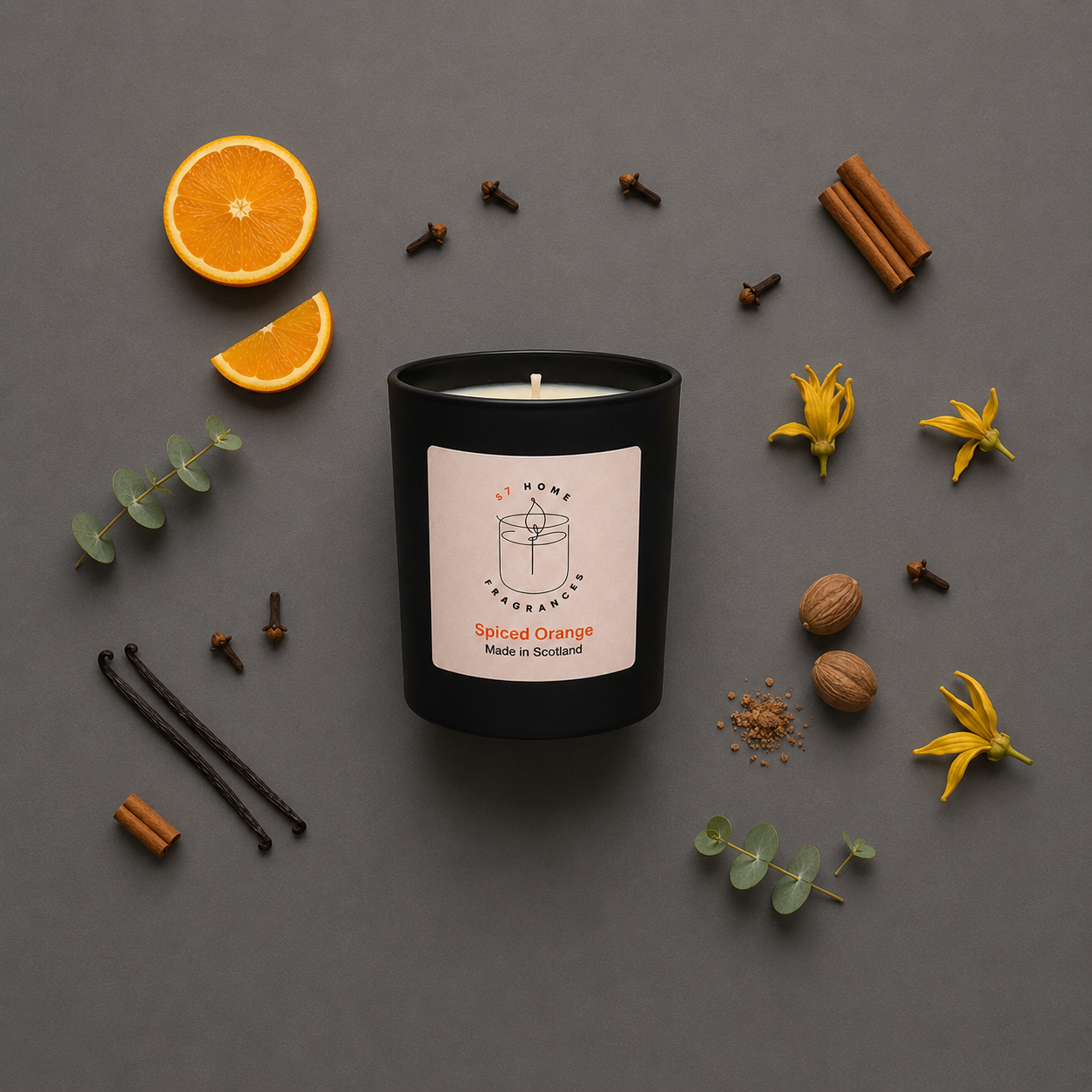 Black candle labeled 'Spiced Orange' surrounded by orange slices, cinnamon sticks, cloves, and flowers on a gray background.